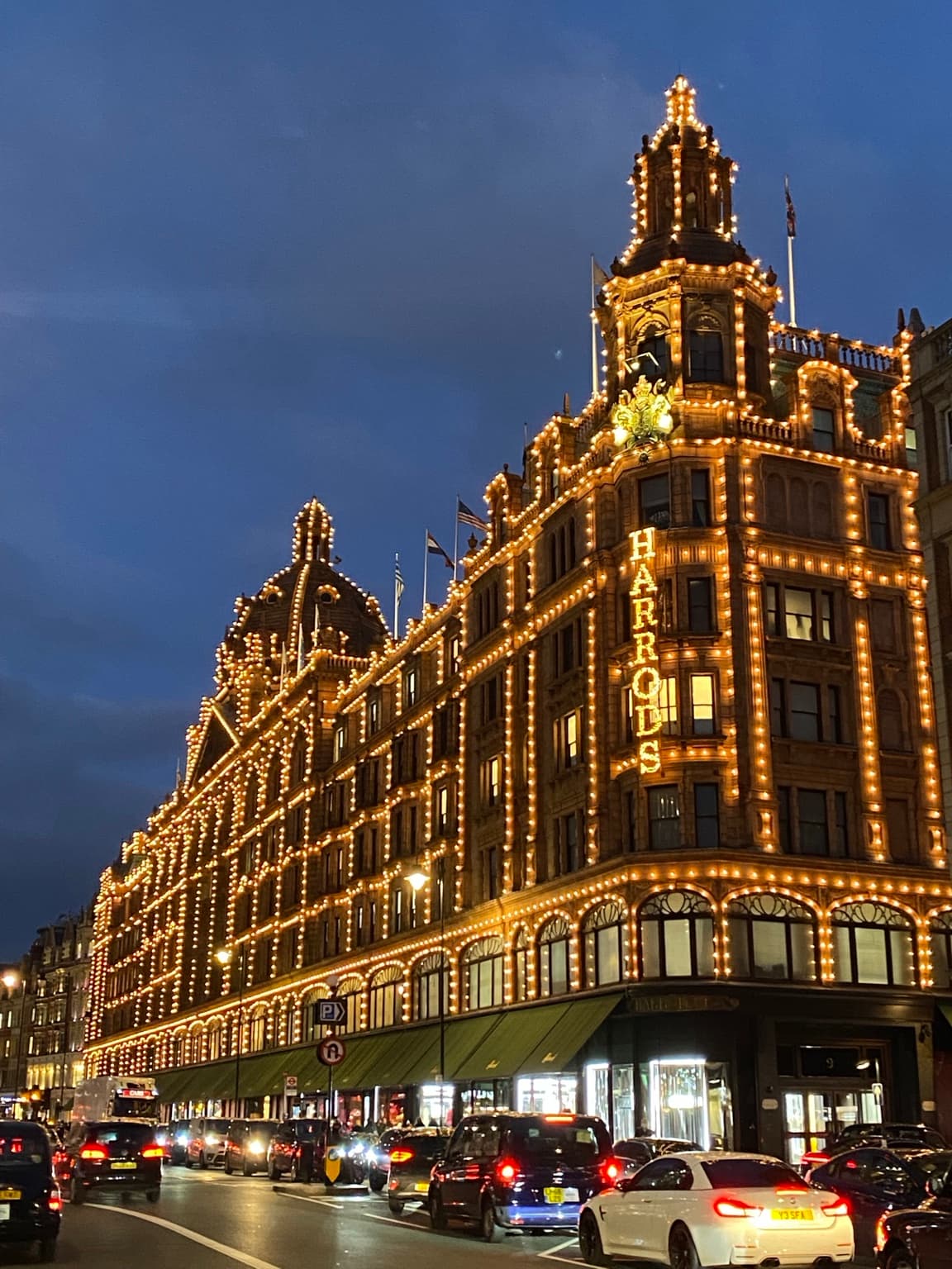 Harrods in Knightsbridge at dusk, with golden lights outlining its towers and façade above passing traffic