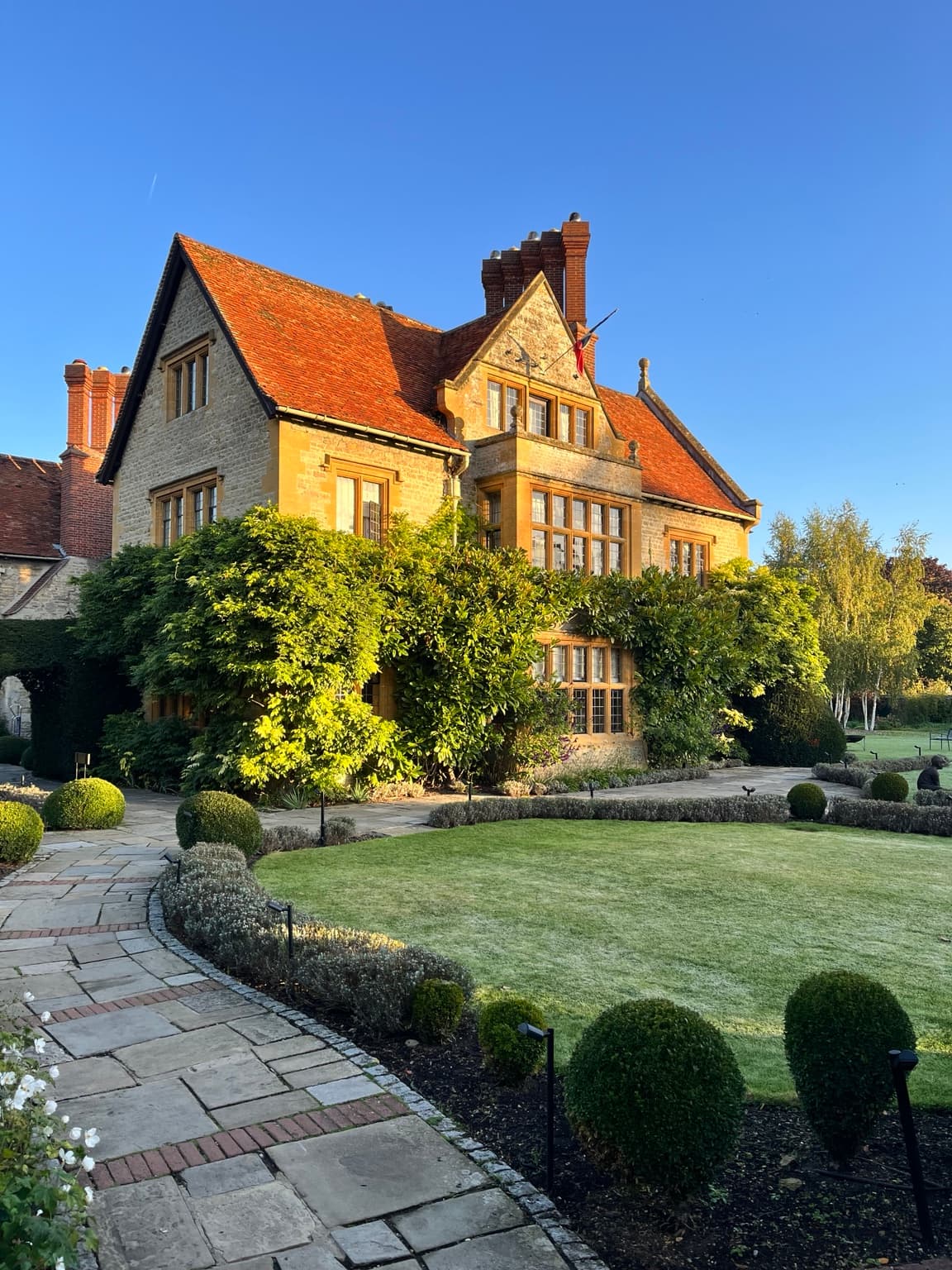 Le Manoir aux Quat’Saisons in golden sunlight, with a stone façade, red-tiled roof and clipped garden borders