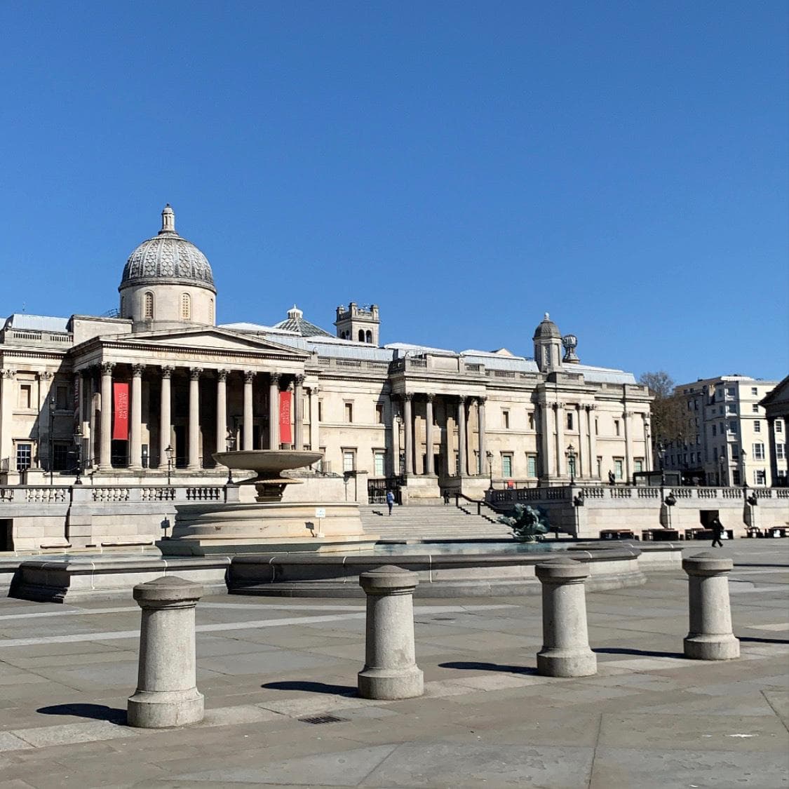 The National Gallery façade and dome above the fountains and stone bollards of Trafalgar Square beneath a clear blue sky